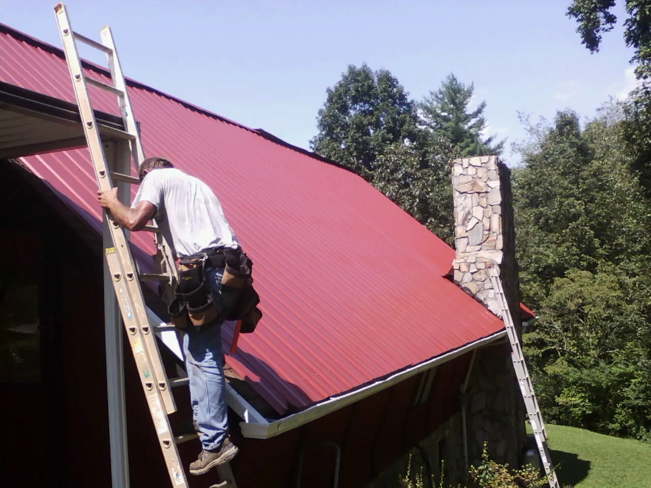 Roofer on a ladder beside a red metal roof and stone chimney — Soffit & Fascia Repair by Roofing Brothers in Terrace Park